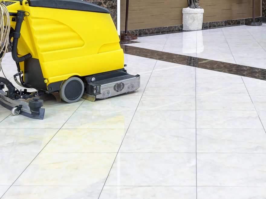 A cleaner in blue overalls guiding a yellow walk-behind floor scrubber across a tiled floor